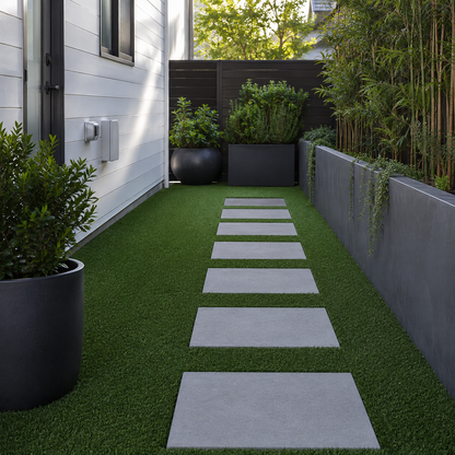 A modern backyard garden pathway made of rectangular light gray stone slabs laid over bright green artificial turf, bordered by a white house wall on one side and tall planters with bamboo and foliage on the other.